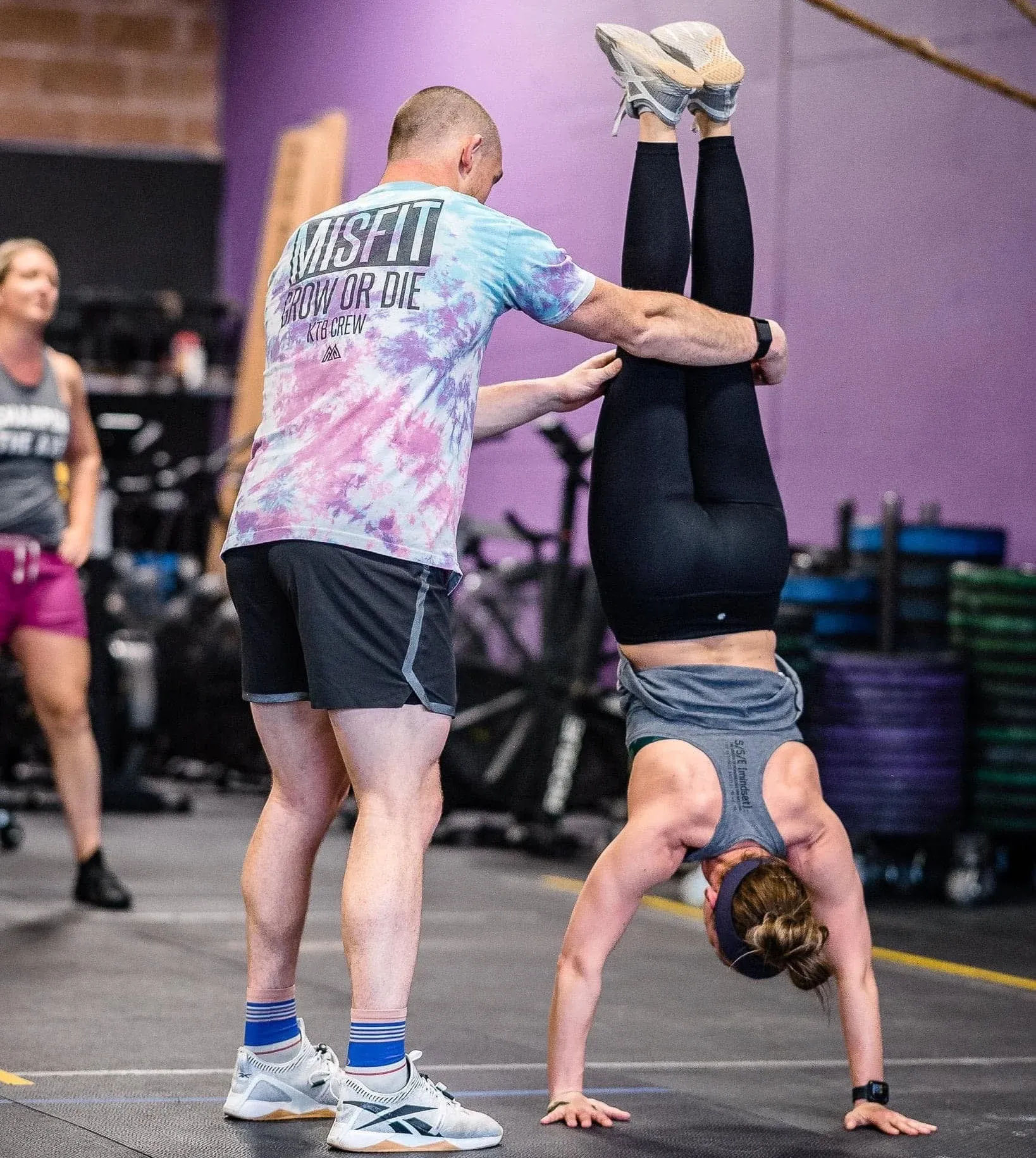 A Misfit Gym Windham coach spots a member's handstand on the gym floor, in front of the purple wall.
