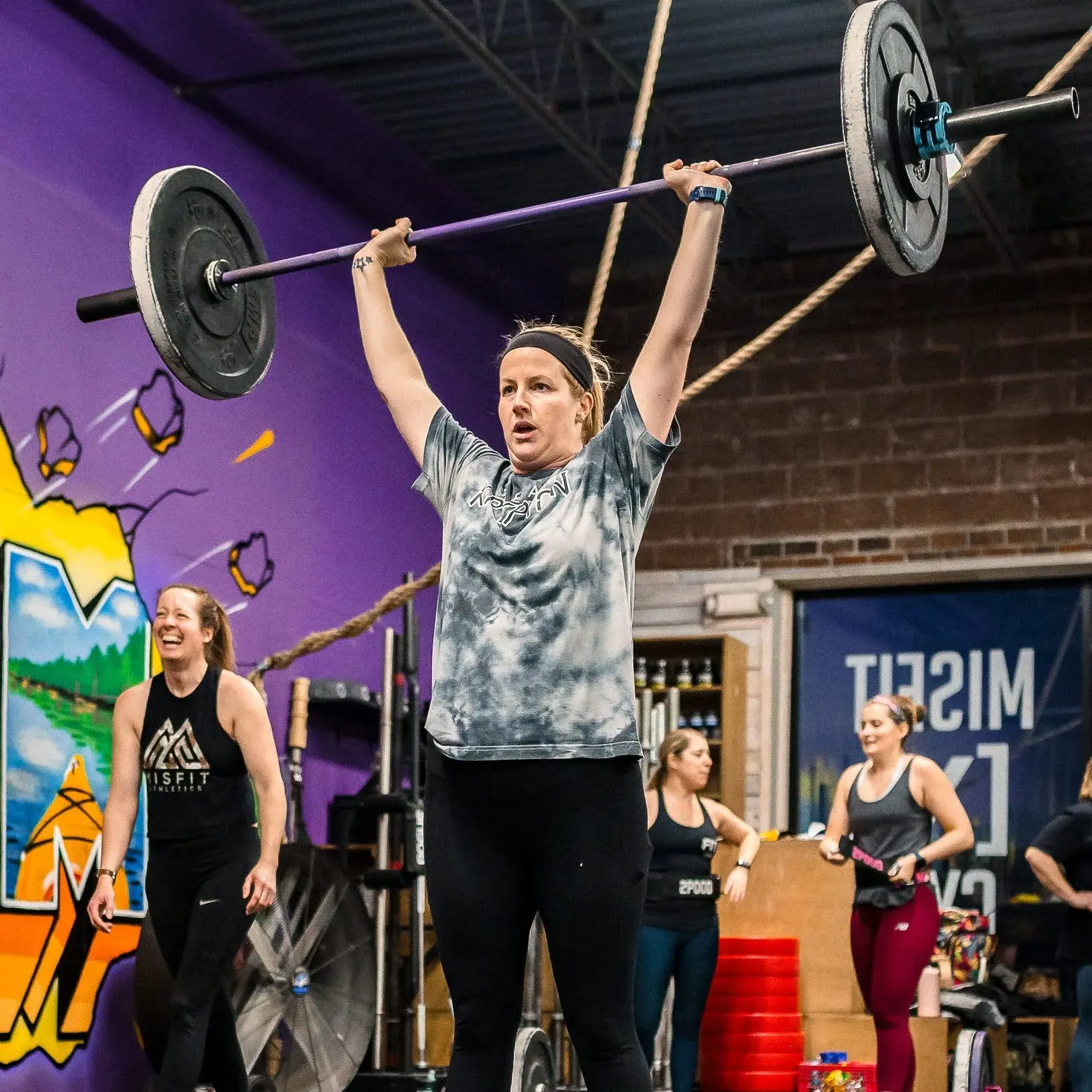 A Misfit Gym Windham member locks out a barbell overhead during class, in front of the gym's purple mural.