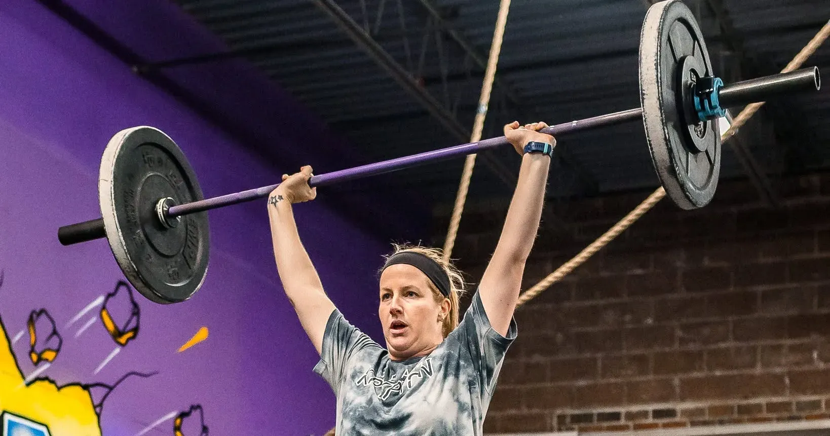 A Misfit Gym Windham member locks out a barbell overhead during class, in front of the gym's purple mural.